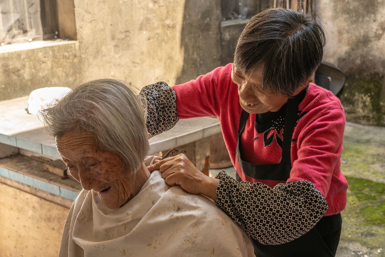 An elderly woman receives a haircut from another woman in a rustic outdoor setting.